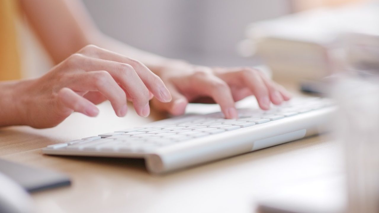 Hands typing on a white keyboard at a sunlit desk