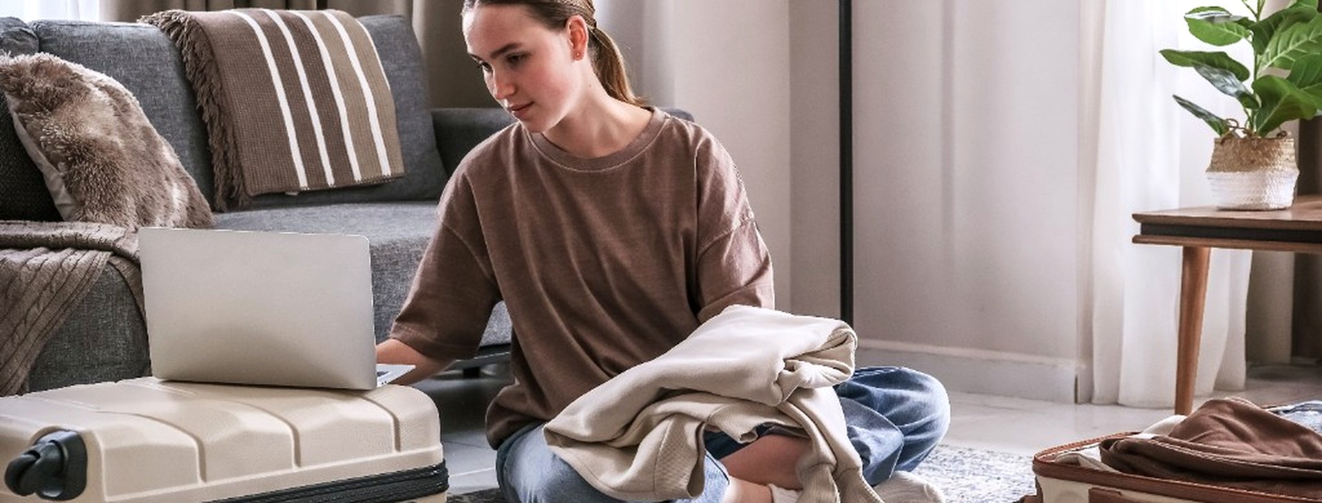 Young woman packing suitcases in a living room with a laptop nearby